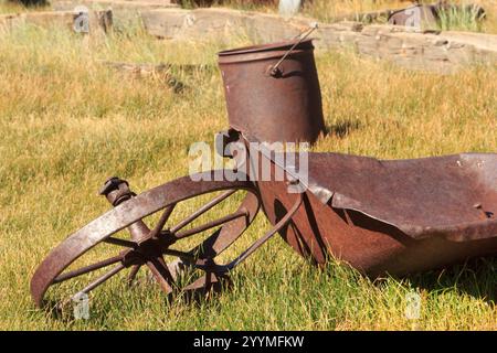 A rusty wheel is laying on the grass next to a broken wheelbarrow. The scene has a sense of abandonment and decay, with the old tools and equipment le Stock Photo