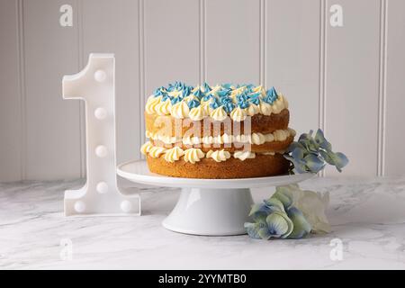 A 1st birthday celebration cake , iced with vanilla buttercream and blue swirls, with a large number one  on the table to the side. Stock Photo