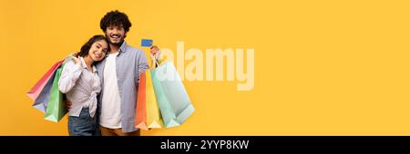 Loving indian couple posing with credit card and colorful shopping bags, making purchases together, yellow background Stock Photo