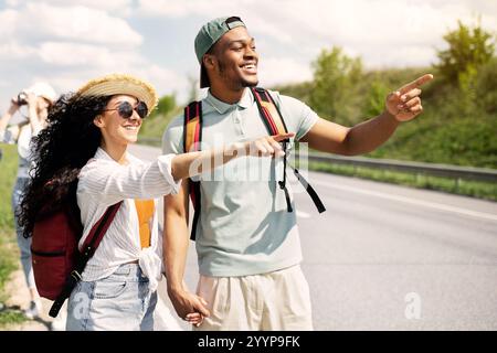 Millennial travelers with backpacks walking along road, hitchhiking in countryside. Group of people having autostop trip Stock Photo