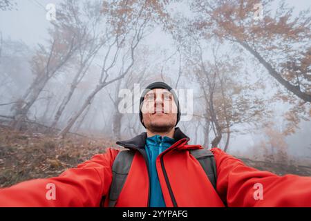 Hiker wearing red jacket taking selfie in foggy forest during autumn Stock Photo
