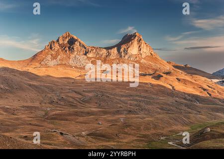 Warm sunlight illuminates the rocky slopes and peaks of two mountains, casting long shadows across the golden valley below Stock Photo