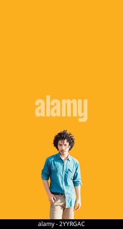 Young man with curly hair wearing a denim shirt and beige pants poses confidently against a vibrant orange background. Minimalistic design with ample Stock Photo