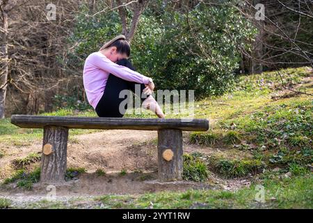 A young woman wearing a pink sweater sits curled up on a rustic wooden bench in a serene outdoor setting. Stock Photo