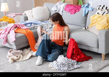 Beautiful young woman with different scattered clothes sitting on floor in messy room Stock Photo