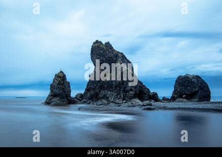 Large jagged rock formations rise dramatically from the calm ocean waters, reflecting a soft blue light as dawn approaches. Gentle waves lap at the sh Stock Photo