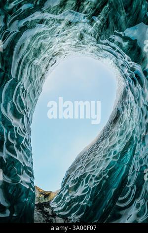 Underneath a frozen cave, curvatures of ice create a beautiful tunnel that frames the vibrant sky above. The surrounding colors reflect the coolness o Stock Photo