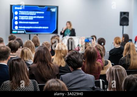 Diverse audience at a seminar with a speaker presenting key information using modern technology. Engaged participants capture moments with smartphones, highlighting interactive learning experience. Stock Photo