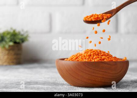 Red lentils pouring from brown wooden spoon into rustic bowl standing on grey kitchen table against white brick wall Stock Photo
