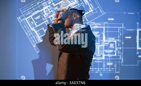 Male architect in hard hat is standing against projector with building plan on it. Stock Photo
