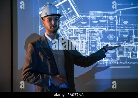 Male architect in hard hat is standing against projector with building plan on it. Stock Photo