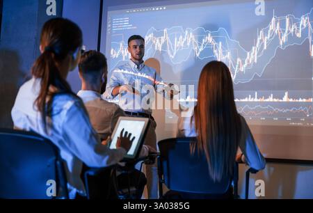 Speaking with audience. Male leader talking to employees, showing the plan on the projector in office of stock exchange company. Stock Photo