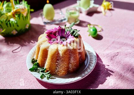 Easter cake decorated with icing sugar and daisy flowers on the table with easter eggs and tulips on  pink tablecloth. Traditional easter cake or swee Stock Photo