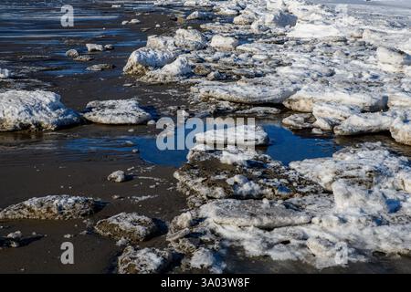 Chunks of broken ice rest on a wet, sandy beach. The ice is dirty. Blue sky reflected in the puddles. Stock Photo
