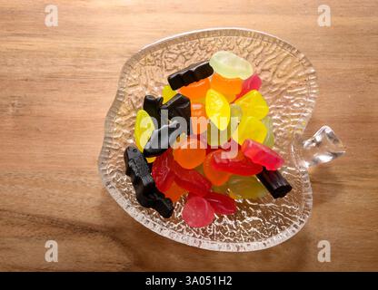 A decorative clear dish holds a vibrant collection of gummy candies in various shapes and colors. The candies are placed on a smooth wooden surface, s Stock Photo
