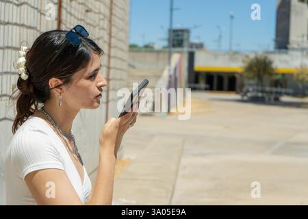 Young latin woman recording a voice message on her phone in a city setting, showcasing modern communication Stock Photo