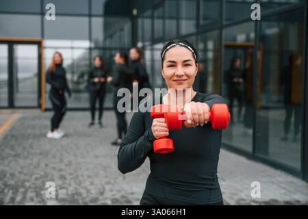 Smiling, doing exercises by using dumbbells. Group of sportive women is outdoors near black building. Stock Photo