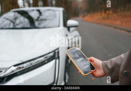With smartphone in hand. Charging process. Woman is standing near her electric car outdoors on the road. Stock Photo