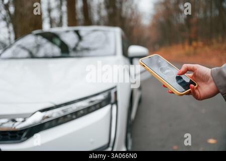 With smartphone in hand. Charging process. Woman is standing near her electric car outdoors on the road. Stock Photo