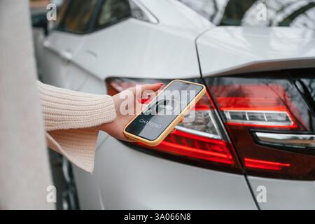 With smartphone in hand. Charging process. Woman is standing near her electric car outdoors on the road. Stock Photo