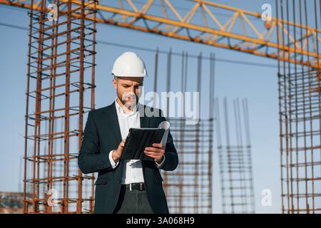 Standing and holding digital tablet. Businessman in formal clothes is on the construction site at daytime. Stock Photo