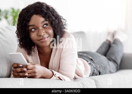 Pensive african woman using smartphone while lying on comfy sofa at home Stock Photo