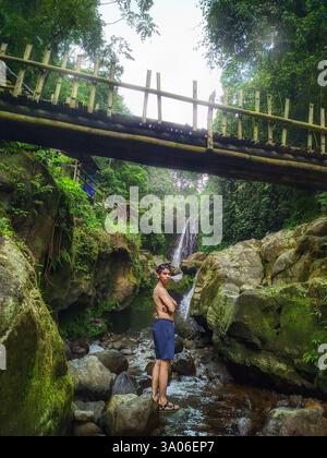 A young man stands among the rocks alongside a flowing stream beneath a small waterfall and rustic wooden bridge, enjoying the tranquil environment of Stock Photo
