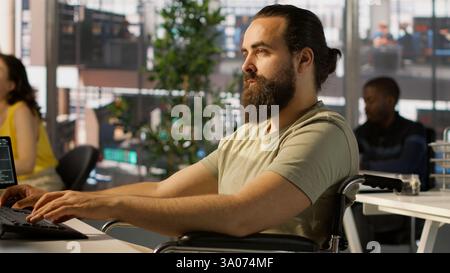 System administrator with disability in inclusive workspace training machine learning models. IT specialist with paraplegia monitoring and maintaining company systems, camera A Stock Photo