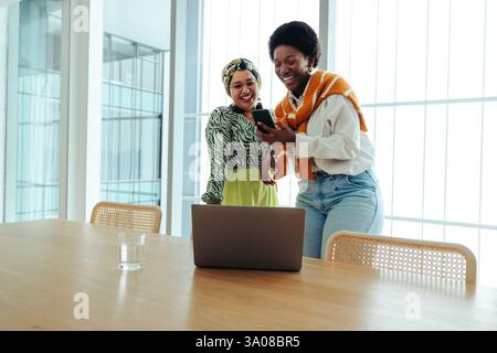 Two successful female business partners engaged in a collaborative moment, laughing and sharing ideas around a laptop. The bright office environment h Stock Photo