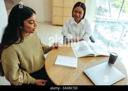 Two friends on campus working together on a project. They are engaged in discussion, with notebooks, a laptop, and coffee on the table. Stock Photo