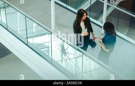 Two businesswomen engage in a friendly conversation while walking through a modern office building. They are discussing work-related topics, reflectin Stock Photo