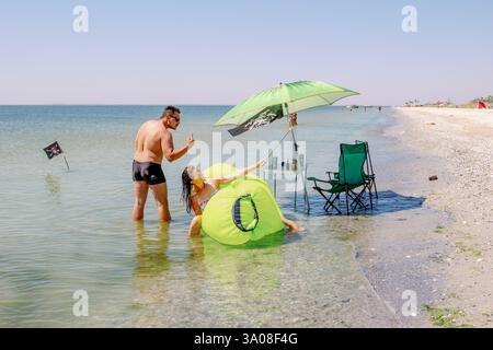 Kinburnska Kosa, Ukraine, August 14, 2019: A couple enjoys a sunny day on the beach taking a selfie on a smartphone next to chairs, a table, a green u Stock Photo
