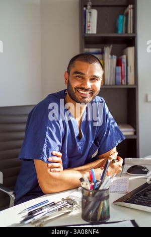 Portrait of smiling male healthcare worker wearing medical scrubs sitting with arms crossed at table in hospital Stock Photo