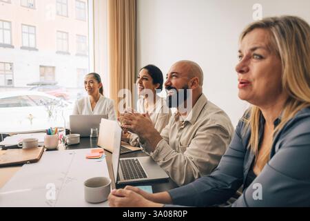 Happy business professional sitting with laptop near female coworkers in meeting room at creative office Stock Photo