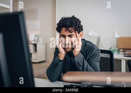 Stressed male entrepreneur touching temples while sitting at desk in creative office Stock Photo