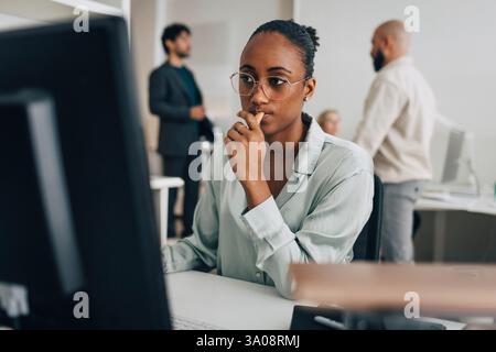 Contemplative young creative professional wearing eyeglasses sitting at desk while working on desktop PC in office Stock Photo