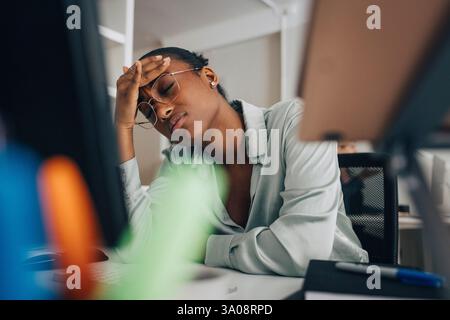 Tired young businesswoman wearing eyeglasses sitting at desk with eyes closed in office Stock Photo