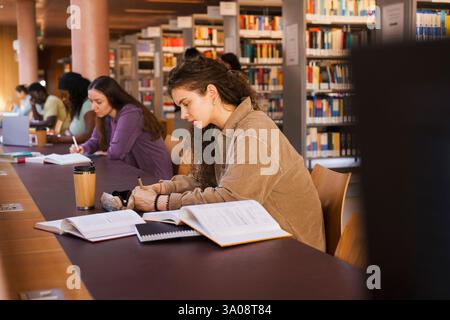 Young female student writing in book while sitting at desk studying inside university library Stock Photo