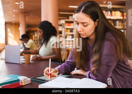 Female student writing assignment on digital tablet while sitting at library desk in university Stock Photo