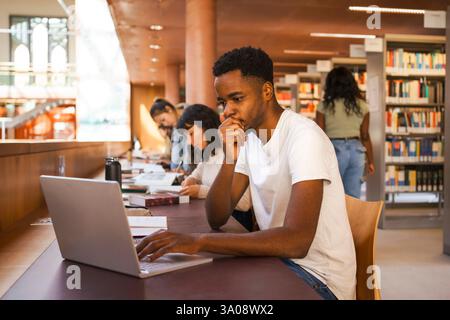 Young male student with hand on chin using laptop while studying in university library Stock Photo