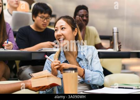 Smiling female student looking at friend holding book while sitting in lecture hall Stock Photo