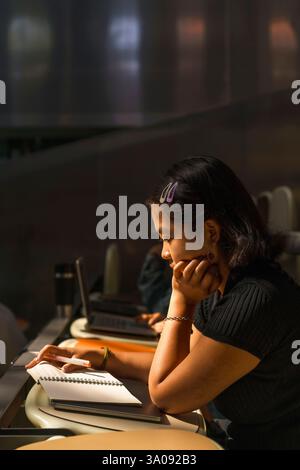 Side view of young female student with hand on chin reading book while sitting in lecture hall at university Stock Photo
