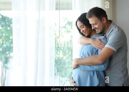 Happy interracial couple enjoying pregnancy together looking at belly at home Stock Photo