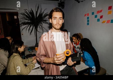 Portrait of young male entrepreneur holding coffee mug and digital tablet with colleagues in background at office Stock Photo