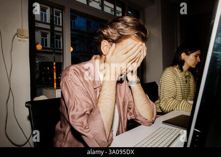 Overworked businessman covering face with hands while sitting at desk in office Stock Photo