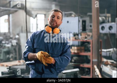 With gloves and protective headphones. Factory worker in blue uniform is indoors. Stock Photo