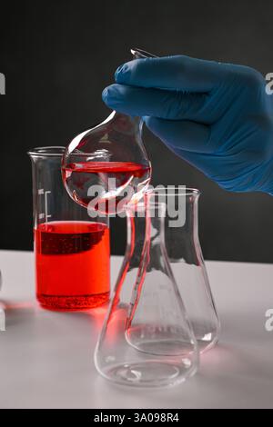 Gloved hand pouring red liquid from laboratory glassware into other beakers and flasks during scientific experiment. Stock Photo