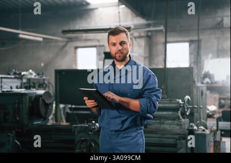 Working by using digital tablet. Man at the factory that is in blue uniform is indoors. Stock Photo
