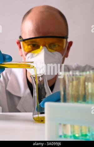 Scientist wearing protective gear pouring yellow liquid from test tube into beaker during experiment in laboratory. Stock Photo