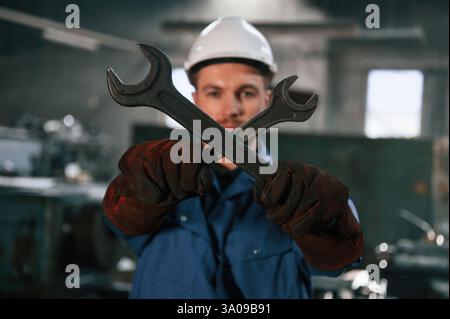 Holding two wrenches. Factory worker in blue uniform is indoors. Stock Photo